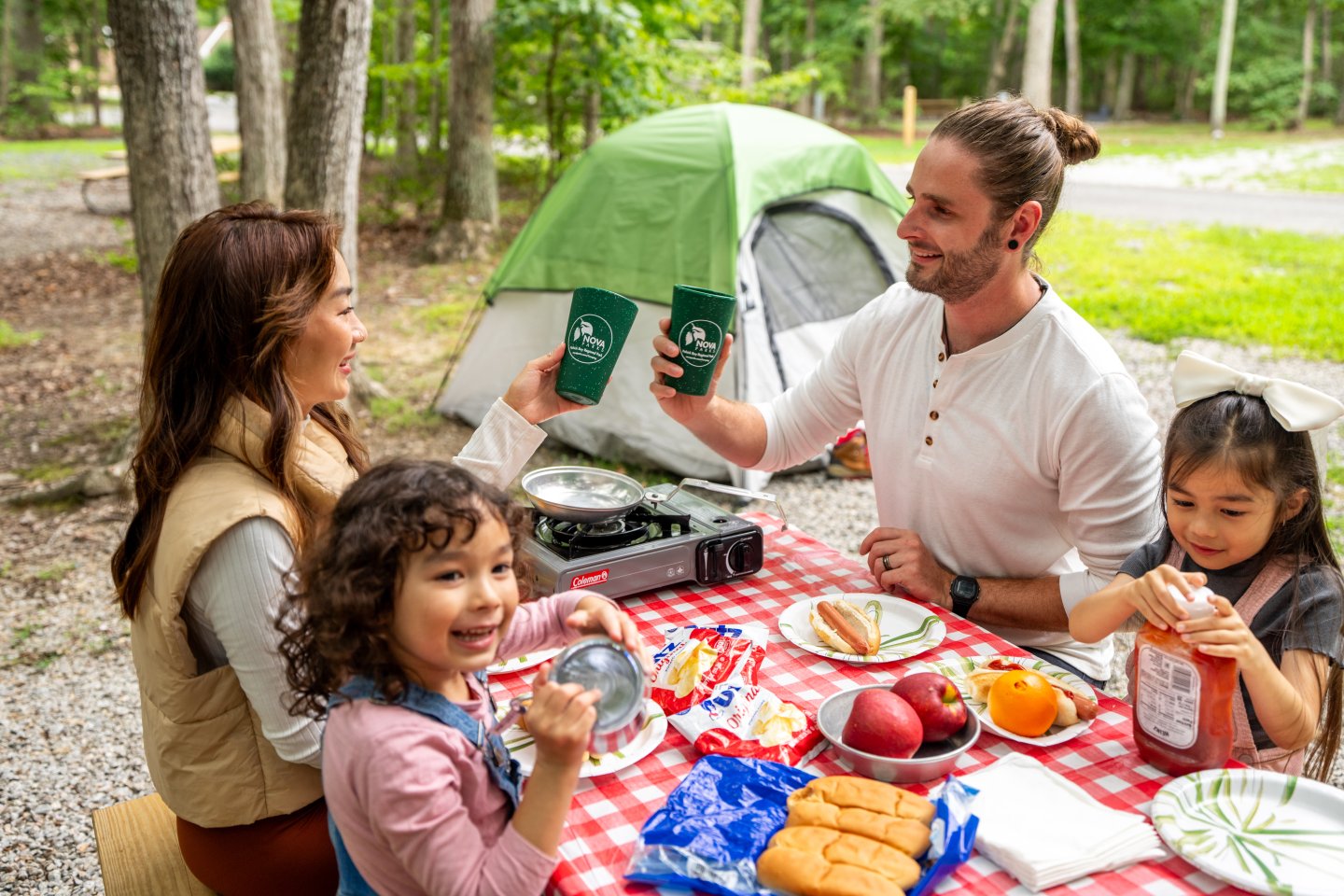 Family enjoying a picnic at a campsite, surrounded by trees and a tent.