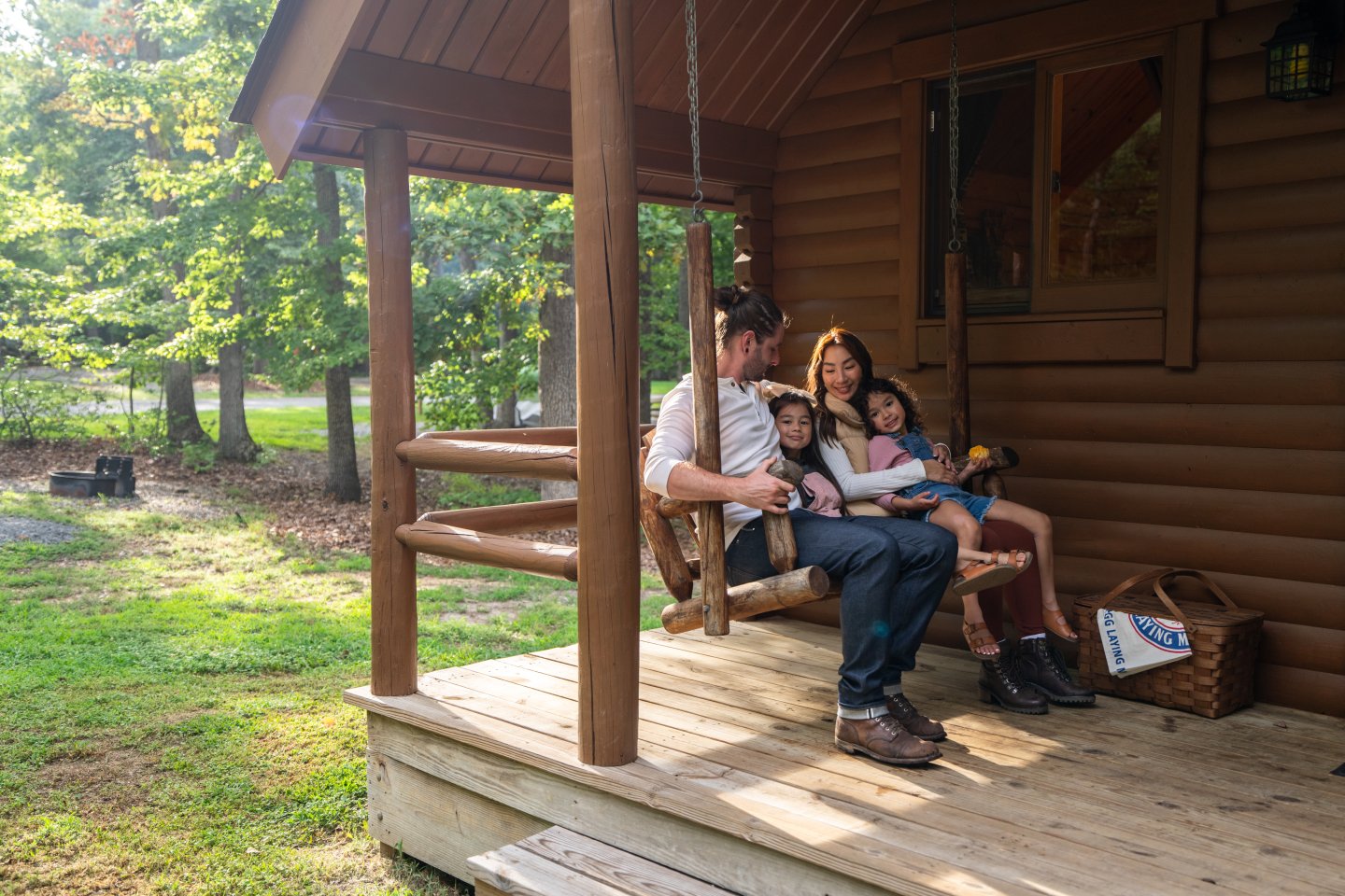 Family sitting on porch swing of a log cabin, surrounded by trees.