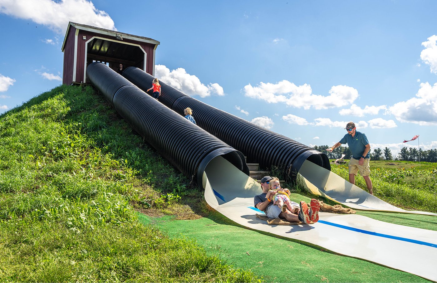 Children sliding down large black tube slides on a grassy hill.