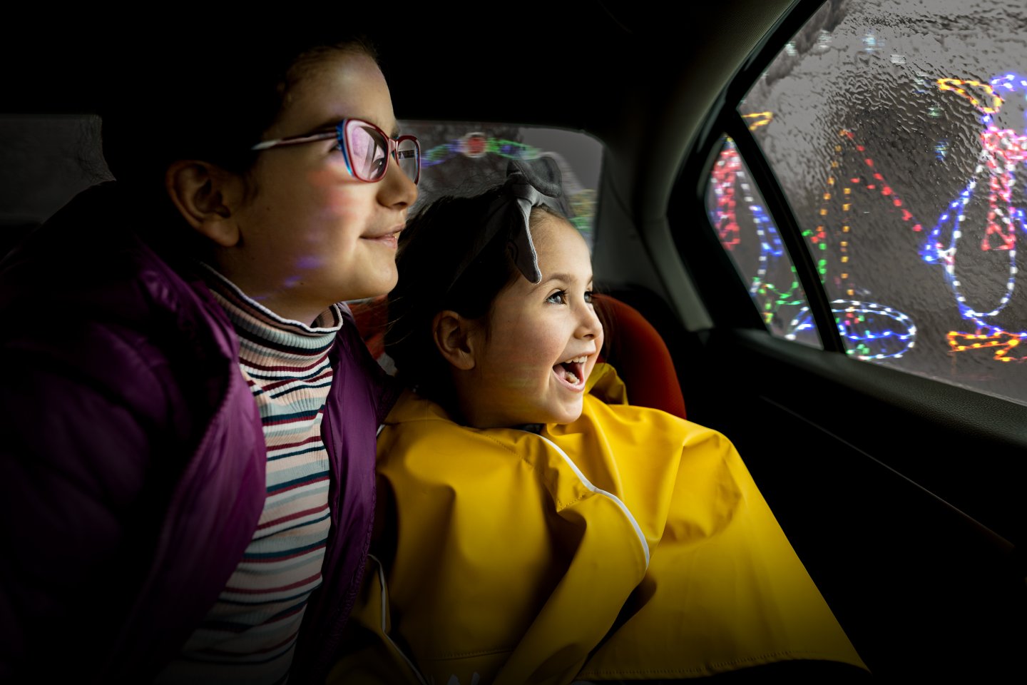 Two children in a car, joyfully watching colorful lights outside the window.