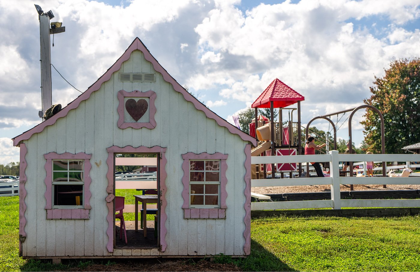 Playhouse with pink trim on grass, playground in the background under cloudy sky.