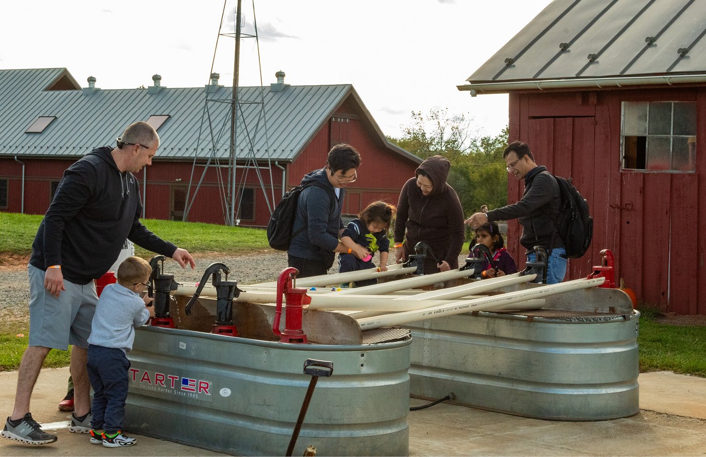People gather around two metal water pumps at a farm setting.