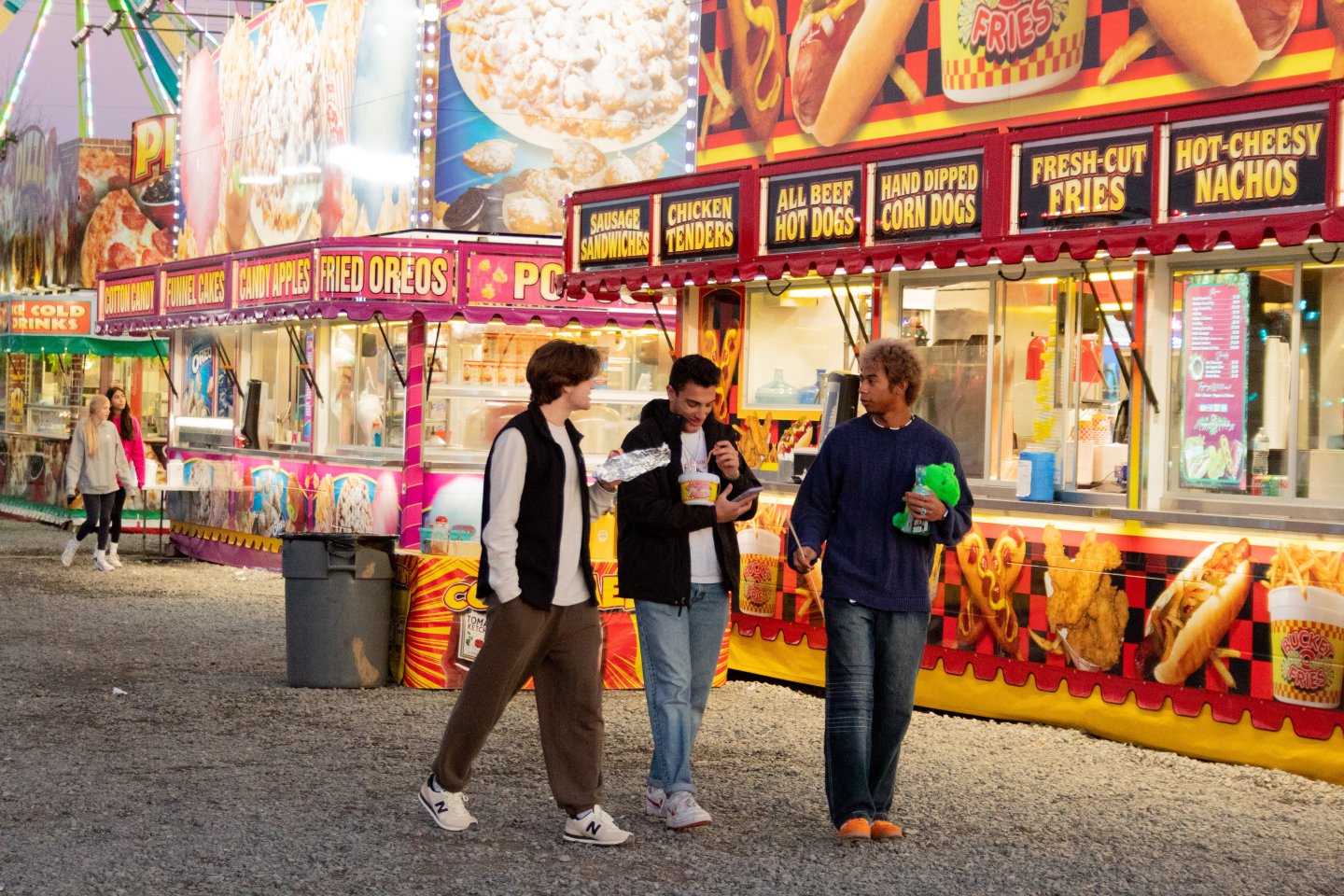 Three friends walk past vibrant carnival food stalls.