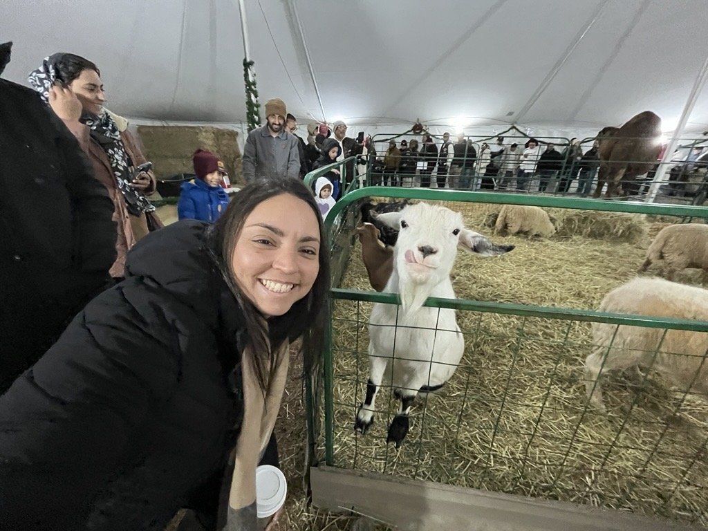 Smiling woman poses next to a goat in a petting zoo under a tent.